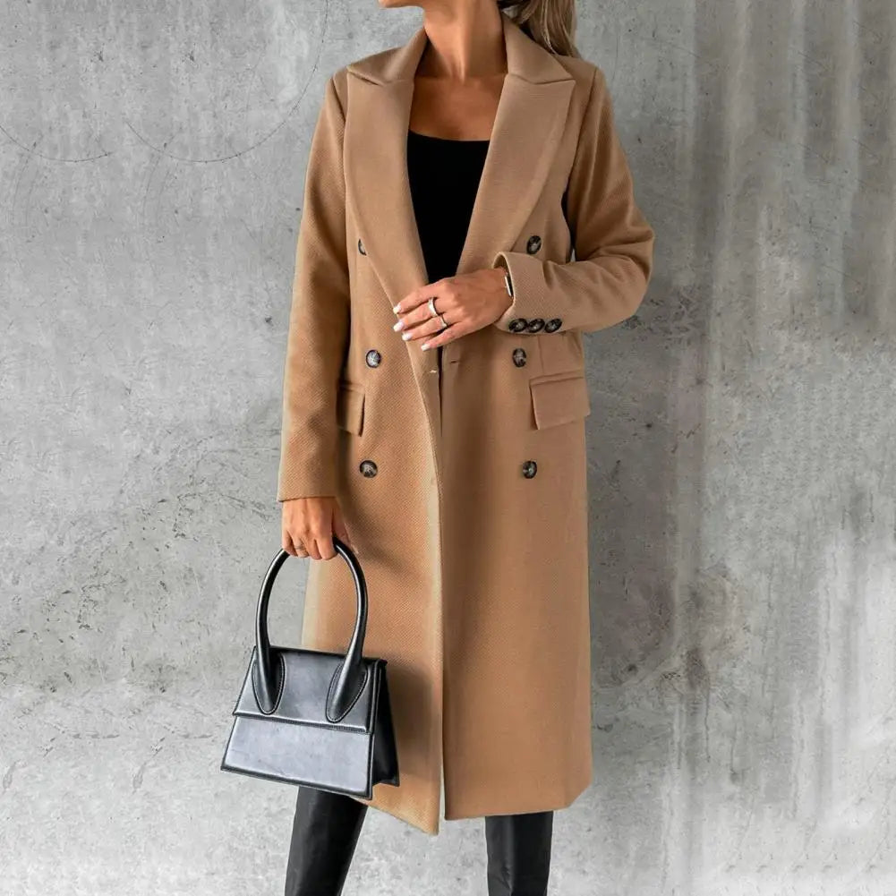 Women's long camel-beige double-breasted trench coat styled over a dark top and black trousers, carrying a black handbag.