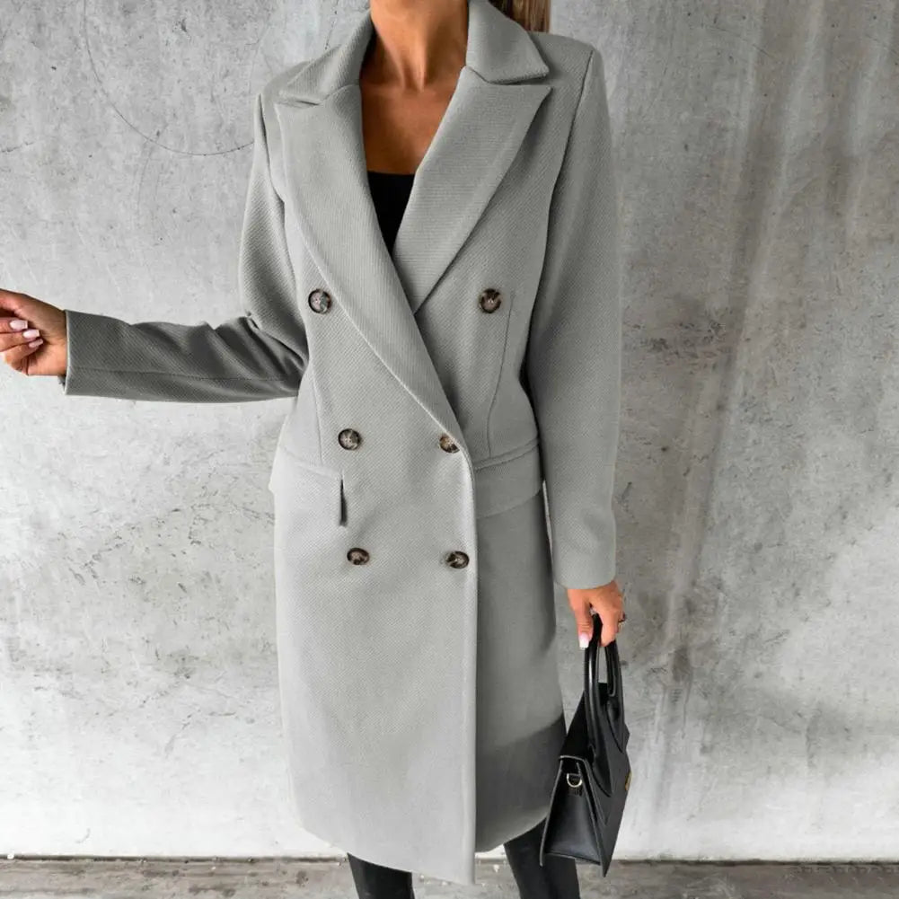 Women's long light grey double-breasted trench coat with flap pockets, styled over a dark top and black trousers, carrying a black handbag.