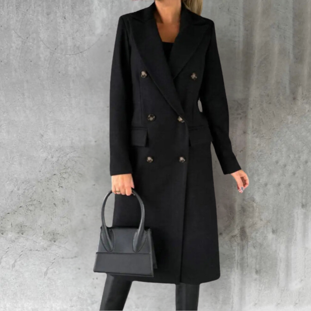 Women's long black double-breasted trench coat styled over a dark top and black trousers, carrying a black handbag.